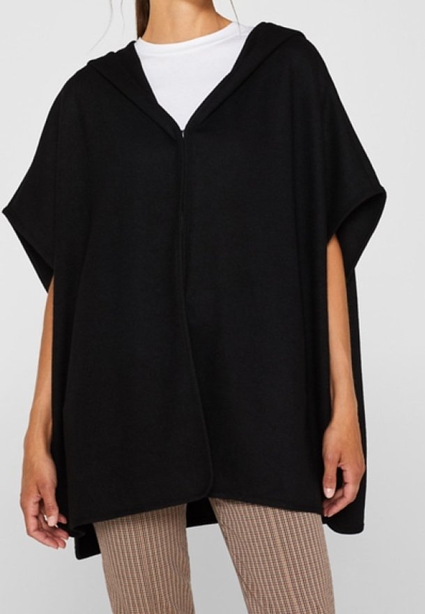 Kobieta w luźnym czarnym ponczo z kapturem, nosząca białą bluzkę i wąskie spodnie w kratę, stoi z luźno opadającymi rękami.