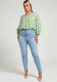 Blusa leggera verde chiaro, trasparente, con motivo floreale e scollo a V; abbinata a jeans in denim azzurro fitted e scarpe con tacco nudo.