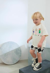 Ljusbeige t-shirt med färggrafik av barn, svart och vit rutiga shorts, och svarta högabottnade sneakers. Släta texturer.