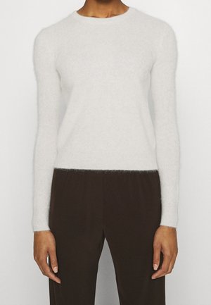 Person trägt einen enganliegenden weißen, flauschigen Pullover mit langen Ärmeln und lockere dunkelbraune Hose, steht vor einem schlichten hellen Hintergrund.