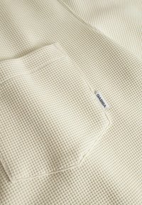 Fehér, gofriknités anyag, téglalap alakú zsebbel és kis márkajellel. Texturált felület, amelyen rácsos minta látható. Puha és könnyű megjelenés.