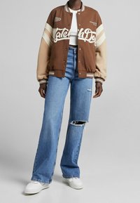 Brun och beige varsityjacka med vit text, matchad med ljusblå slitna byxor med vida ben och vita sneakers.