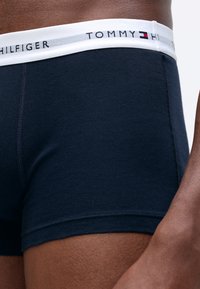 Tuvumā redzami tumši zili bokseršorti ar baltu jostu, uz kuras redzama "Tommy Hilfiger" zīmola apzīmējums, ko valkā persona ar tumšu ādu.