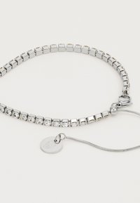 Sølvfarget tennisarmbånd med klare kvadratiske krystaller og en liten rund anheng på hvit bakgrunn.