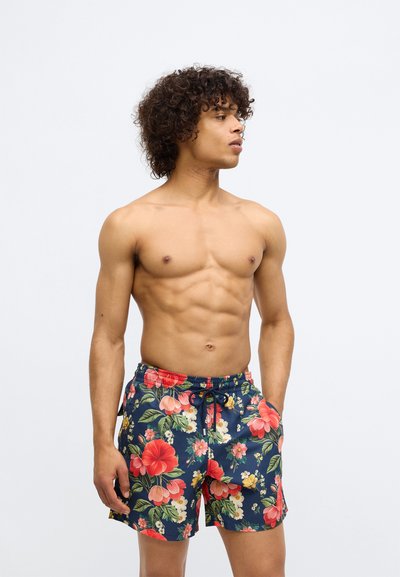Homme torse nu avec des cheveux bouclés portant un short de bain bleu marine avec un imprimé floral rouge, rose et blanc, se tenant avec une main dans la poche.