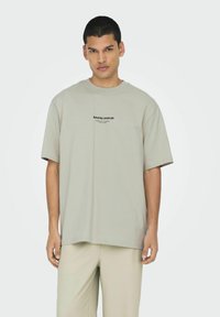 Lichtgrijze oversized T-shirt met korte mouwen, voorzien van een zwart gedrukt logo op de voorkant. De stof lijkt glad en zacht van structuur.