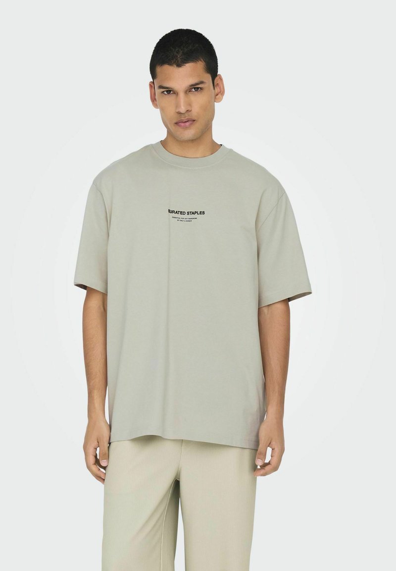 Lichtgrijze oversized T-shirt met korte mouwen, voorzien van een zwart gedrukt logo op de voorkant. De stof lijkt glad en zacht van structuur.