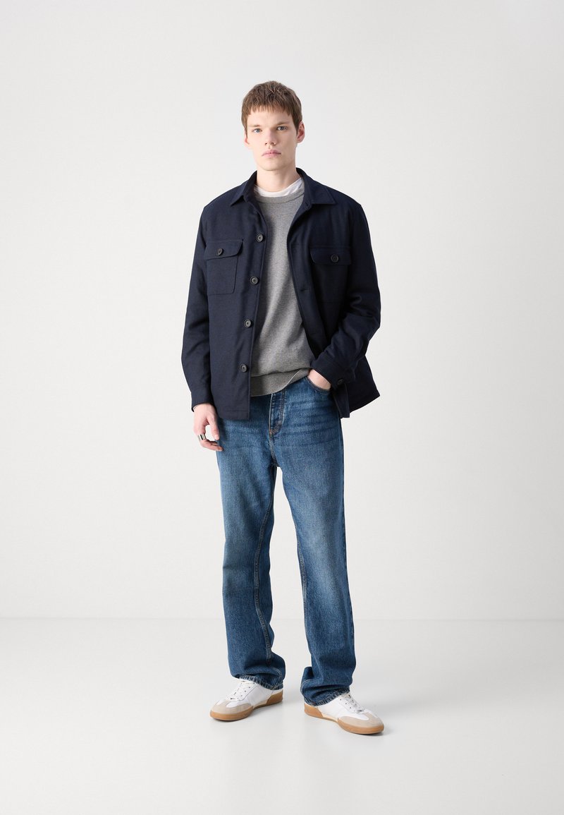 Veste marine à boutons montée avec deux poches poitrine, pull gris en dessous, jean bleu et baskets blanches avec semelles en gomme, se tenant sur un fond blanc.