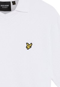 Vit polo skjorta med texturerad tyg, V-hals och en liten svart- och gul örnlogotyp på vänster bröst. Avslappnad passform.