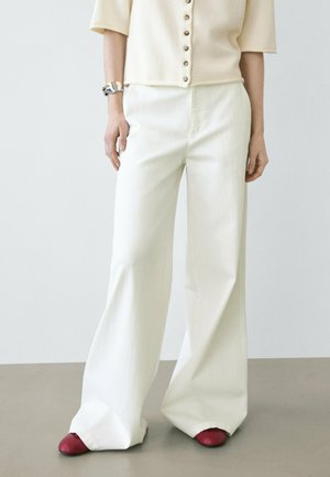 Donna che indossa pantaloni bianchi a gamba larga, top crema a maniche corte con bottoni, braccialetto argentato e scarpe rosse, in piedi su pavimento grigio.