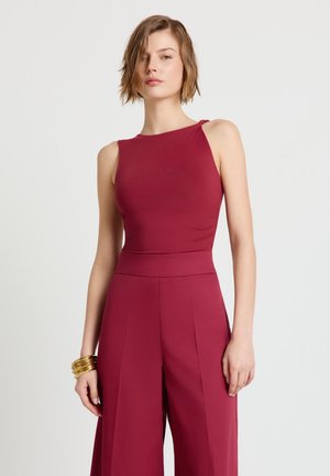 Donna che indossa una tuta senza maniche color bordeaux con pantaloni a gamba larga e braccialetti d'oro al polso sinistro, in piedi contro uno sfondo chiaro e semplice.