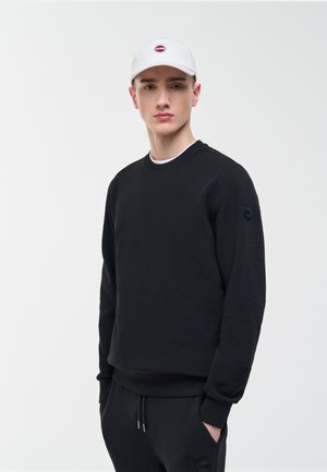 Młody mężczyzna ubrany w czarną bluzę i spodnie z rękami w kieszeniach, w białą czapkę z daszkiem z czerwonym logo na jednolitym tle.