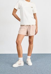 Persona che indossa sneaker Adidas bianche, calze bianche, pantaloni corti beige e una maglietta bianca con una piccola grafica sul petto, in piedi su un tappeto blu.