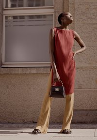 Vestido plissado vermelho combinado com calças plissadas amarelas. Modelo segura uma pequena bolsa preta. O fundo apresenta uma parede de pedra texturizada e uma janela.