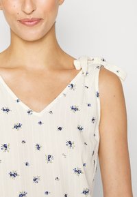 Vestido sem mangas de creme claro com decote em V e padrão floral em azul e verde. Apresenta alças amarradas e um tecido texturizado.