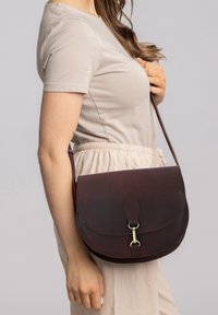 Sac à bandoulière en cuir marron avec fermeture à rabat, forme incurvée et détails en laiton. Texture lisse, porté avec une tenue beige clair.