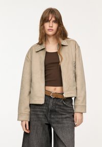 Giacca in suede beige con colletto borchiato e chiusura con zip, abbinata a una maglietta marrone e jeans a gamba larga grigi. Texture liscia, vestibilità comoda.