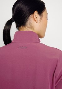 Polo de color púrpura con un collar alto, que presenta un tejido texturizado y un discreto bordado "COLD RDY" en la parte posterior.