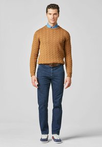 Pull en maille torsadée couleur camel, chemise bleue en dessous, jean foncé et baskets basses à semelle blanche avec des accents bleu marine.