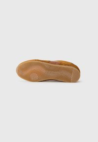 Bruine suède schoen met een gestructureerde rubberen zool met een golvend patroon, gestikte accenten en een cirkelvormig logo op de hiel van de zool.