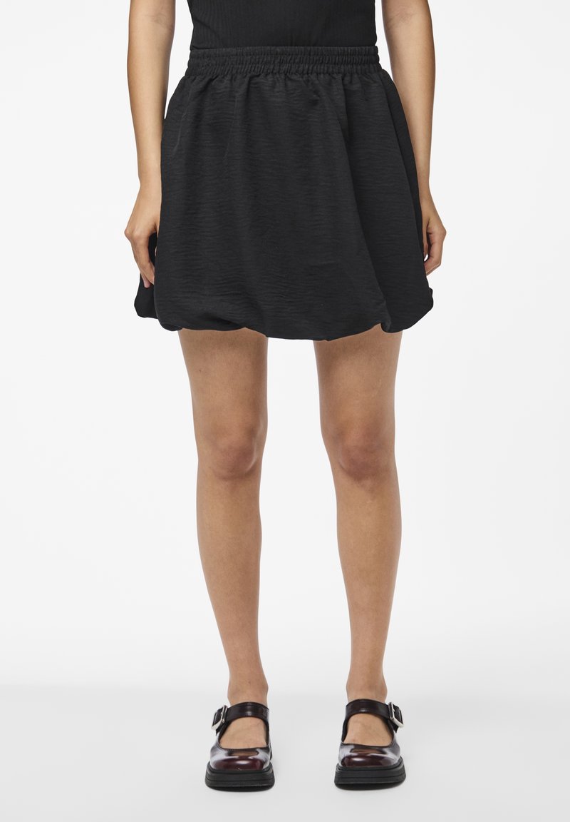 Černá sukně s texturovaným, lehkým materiálem. Obsahuje elastický pas a rozšířený tvar nad kolena. Spárována s tmavými botami.