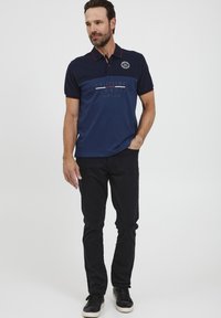 Sötétkék férfi pólóing kontrasztos gallérral, rövid ujjakkal és logó részlettel; slim fit fekete nadrág és kényelmes fekete sportcipő.