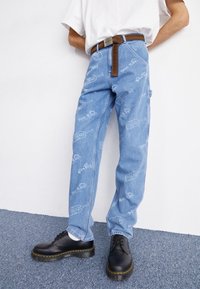 Hellblaue Denim-Jeans mit gedruckten Angel-Motiven, gerades Bein-Design und braunem Ledergürtel. Kombiniert mit schwarzen Schnürschuhen.
