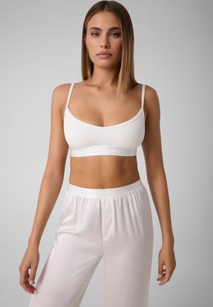 Femme aux cheveux châtain clair et raides portant un bralette blanc côtelé et un pantalon de pyjama en satin blanc sur un fond gris uni.