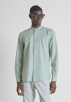 Mann trägt hellgrünes Hemd mit Mandarinkragen und weiße Hose, steht mit entspannten Händen, trägt große schwarze Sonnenbrille, schaut nach rechts.