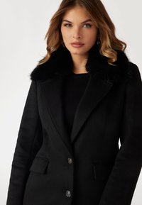 Cappotto in misto lana nero con colletto in pelliccia, chiusura frontale doppiopetto con bottoni decorativi e due tasche laterali.