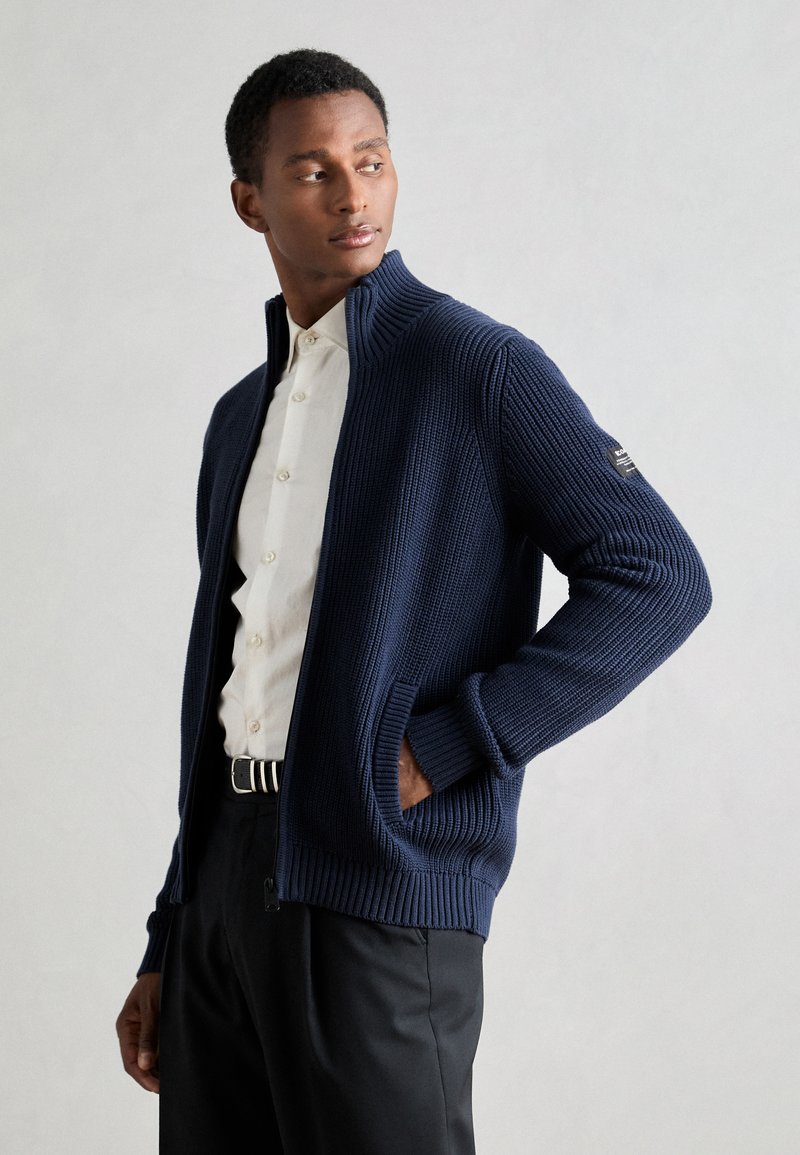 Maglione a zip blu navy con texture a coste, colletto alto e tasche laterali, abbinato a una camicia bianca con bottoni e pantaloni neri.