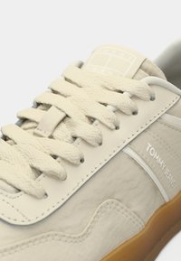 Gros plan sur une sneaker beige Tommy Jeans avec des lacets texturés, des détails cousus et une semelle en gomme, mettant en avant les logos de la marque sur la languette et sur le côté.