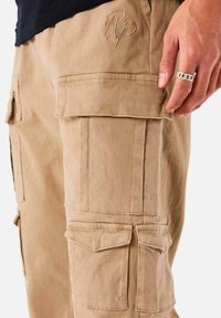 Beige cargo bukser med flere lommer, afslappet pasform og broderet logo-detail på låret. Struktureret bomuldsstof.