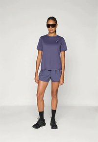 Set da t-shirt e pantaloni corti sportivi viola, realizzato in tessuto leggero, con un logo discreto. Gli accessori neri includono occhiali da sole e scarpe.