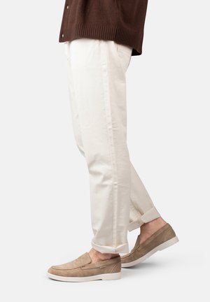 Persoon die beige suède instapschoenen draagt en opgerolde witte broek, staand tegen een effen lichte achtergrond.