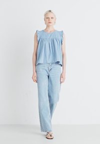 Svetlo modra denim brezrokavnik s kerbljasto obrobo ramen, v kombinaciji s svetlo modrimi širokimi džinsami. Minimalističen dizajn, gladka tekstura.