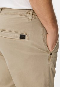 Beige Baumwoll-Chinos mit seitlichen und hinteren Taschen. Sie verfügen über ein kleines schwarzes Markenetikett auf der Hintertasche und eine glatte Textur.