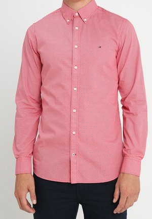 Muž nosící přiléhavou košili s dlouhým rukávem na knoflíky s malým geometrickým vzorem v červené a bílé barvě.