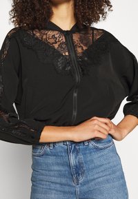 Black bomber jacket featuring floral lace accents on the shoulders and front. Soft fabric, zip closure, and relaxed fit. Worn with blue denim.