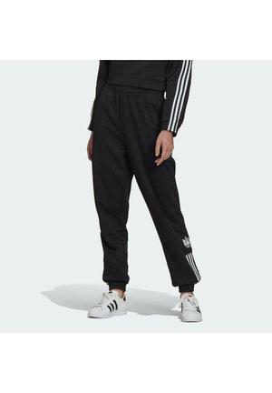 Persoon draagt zwarte Adidas trainingsbroek met witte strepen en logo, gecombineerd met witte Adidas sneakers met zwarte strepen.