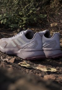 Hellgraue Adidas Terrex Trailrunning-Schuhe auf einem felsigen, mit Blättern bedeckten Außenboden mit grünen Pflanzen im Hintergrund.