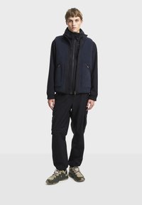 Noćno plava flis prsluk preko crne jakne na zatvaranje s patentnim zatvaračem, uparen s crnim cargo hlačama i smeškastozelenim patikama, s omogućnim džepovima i opuštenim krojem.