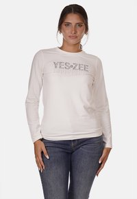 T-shirt bianco a maniche lunghe con un logo metallico "YES-ZEE" sul petto. Tessuto morbido con una silhouette aderente. Indossato con jeans in denim scuro.