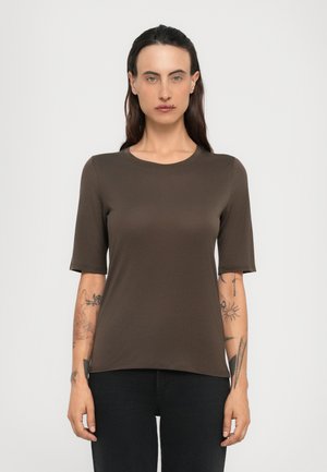 Kvinne med langt mørkt hår iført en brun kortermet skjorte og svarte bukser, som viser tatoveringer på begge underarmer mot en ensfarget bakgrunn.
