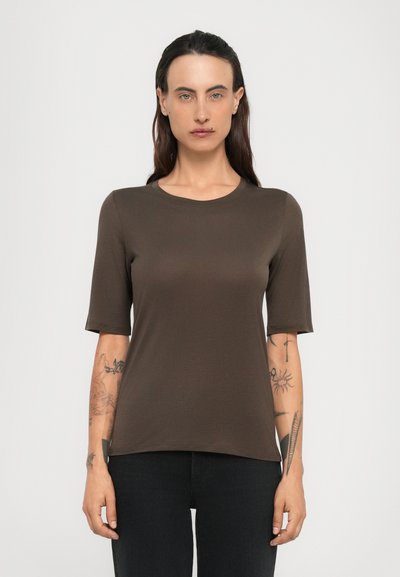 Donna con lunghi capelli scuri indossa una maglietta marrone a maniche corte e pantaloni neri, mostrando tatuaggi su entrambi i avambracci contro uno sfondo semplice.