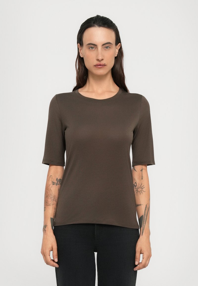 Donna con lunghi capelli scuri indossa una maglietta marrone a maniche corte e pantaloni neri, mostrando tatuaggi su entrambi i avambracci contro uno sfondo semplice.