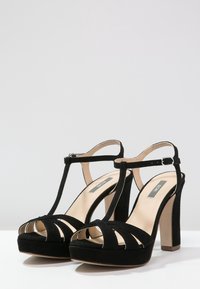 Czarne zamszowe sandały na wysokim obcasie z designem T-strap, odkrytymi palcami i grubym obcasem. Posiadają zapięcie na kostce oraz platformę.