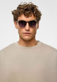 Ronde zwarte zonnebril met gradient lenzen. Het montuur heeft zilveren accenten. Draagt een lichtbeige sweater met ronde hals. Gevoelige huid zichtbaar.