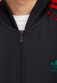 Svart zip-up träningsoverallsjacka med röda ränder och grön logotyp på vänster bröst. Slät textur, hög krage och svarta dragkedjedetaljer.