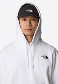 Sudadera blanca con capucha, cordones y logotipo negro en el lado izquierdo del pecho. Llevada con una gorra negra que también muestra el logotipo.